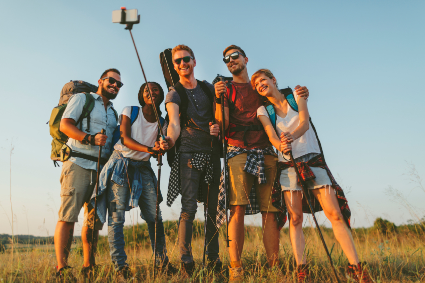 Group of young hikers making group selfie with smart phone and selfie stick. They enjoy sunny day and having fun. Male in the middle holding selfie stick. All of then looking at camera and smiling, and holding hiking poles in hands. Males are wearing sunglasses.