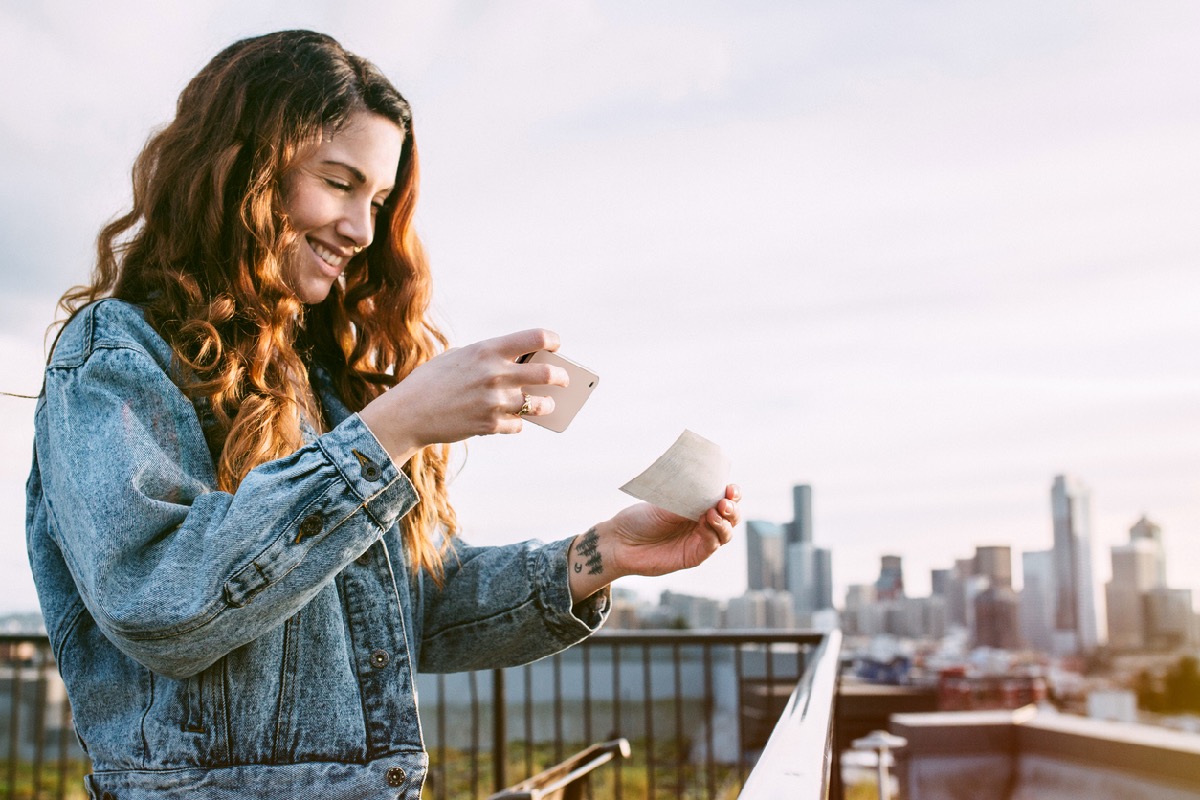 A beautiful Caucasian adult woman on a Seattle roof top taking a picture of a check with her smart phone for a Remote Deposit Capture.  She smiles, wearing modern stylish clothing.  Seattle cityscape on the skyline in background.