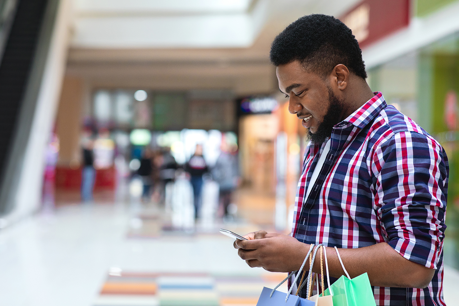 App With Discounts. Smiling Black Man With Shopping Bags Using Smartphone In Mall, Checking Sale Deals, Side View With Free Space