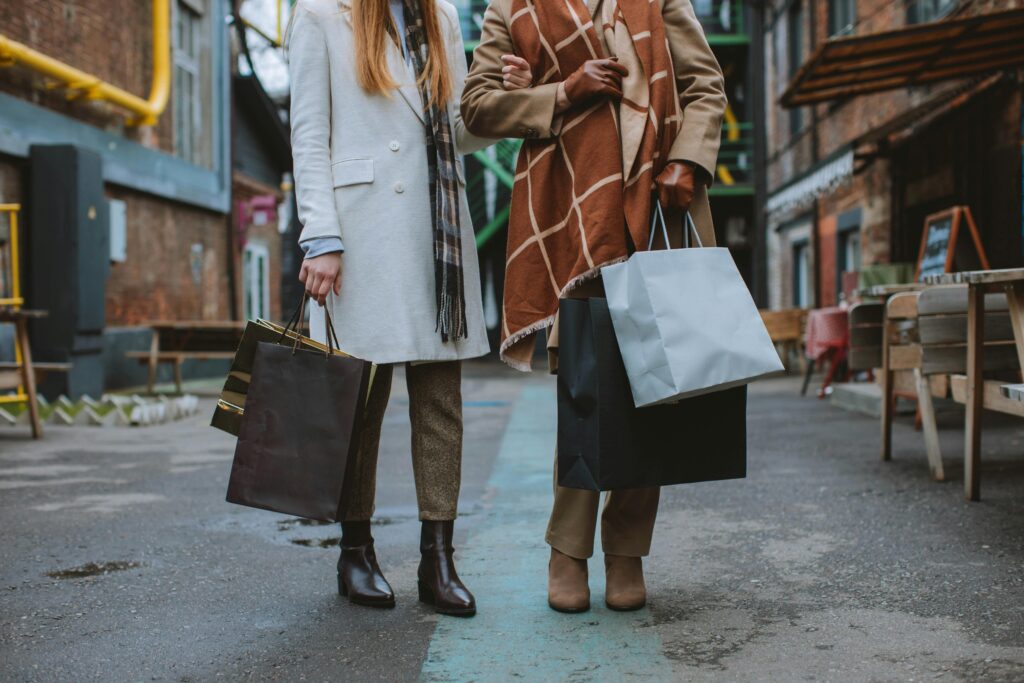 Two women in coats walking down an urban alley, carrying multiple shopping bags in each hand.