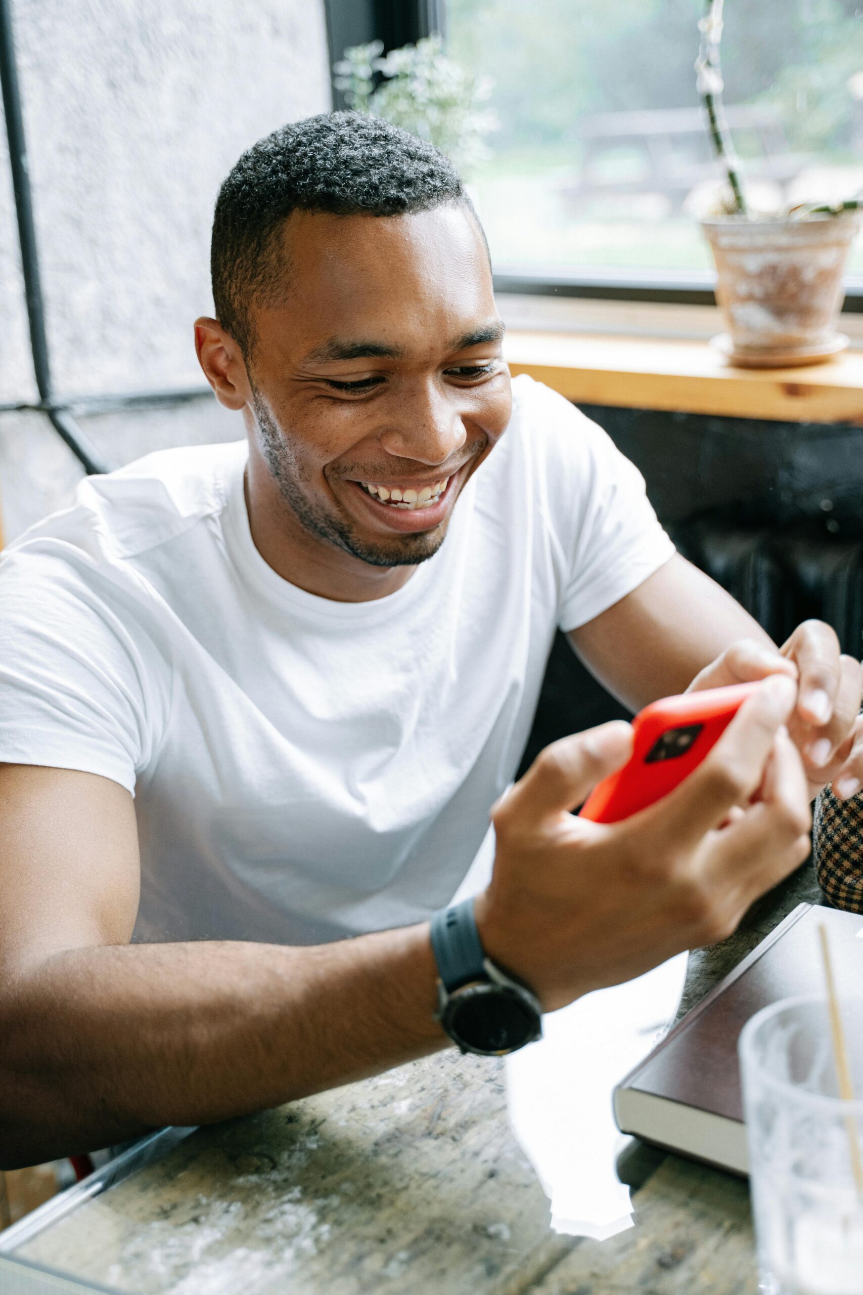 Man in a white T-shirt smiles while looking at a red smartphone at a cafe table by a window with a potted plant nearby