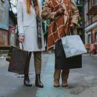 Two women in coats walking down an urban alley, carrying multiple shopping bags in each hand.