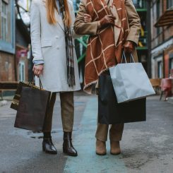 Two women in coats walking down an urban alley, carrying multiple shopping bags in each hand.