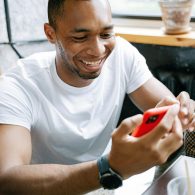 Man in a white T-shirt smiles while looking at a red smartphone at a cafe table by a window with a potted plant nearby