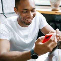 Man in a white T-shirt smiles while looking at a red smartphone at a cafe table by a window with a potted plant nearby